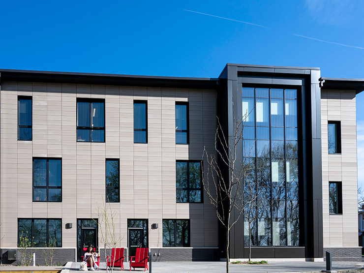 A student sits outdoors in a red chair with a modern building, with lots of open windows, behind them.