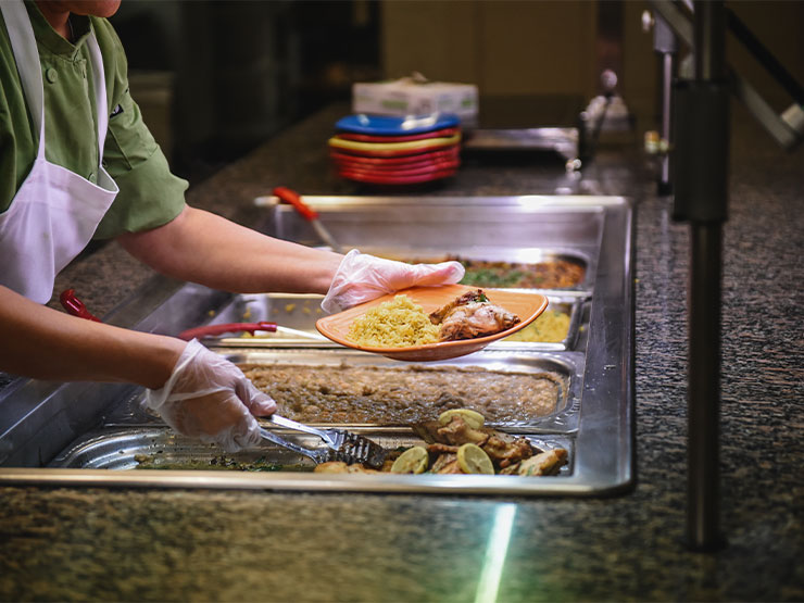 Two gloved hands serve food on a plate in a cafeteria setting.