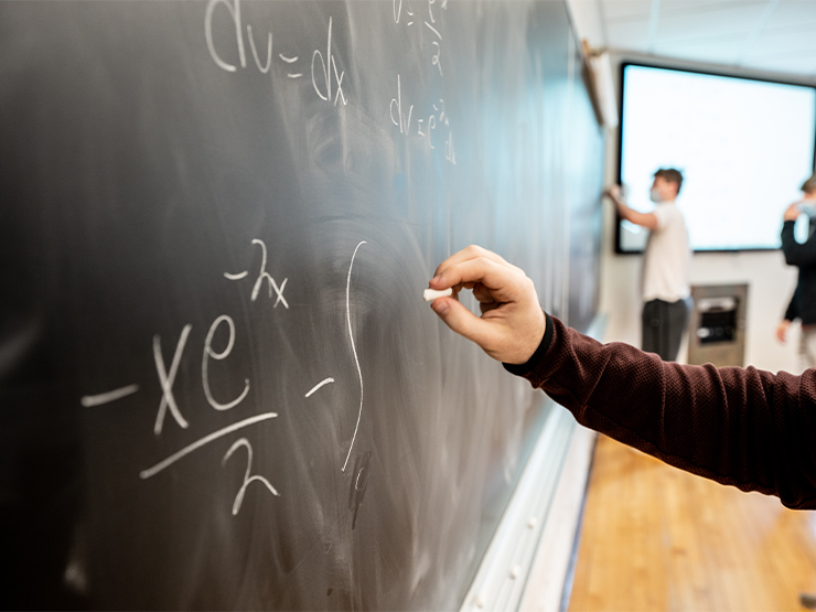 A hand is extended toward a chalkboard where part of a formula is written.