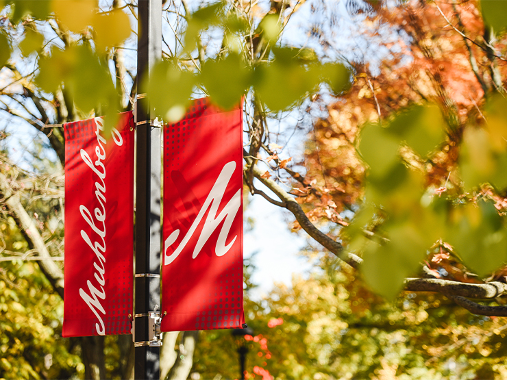 Two red Muhlenberg flags hang off a light pole on campus, framed by colorful fall foliage.