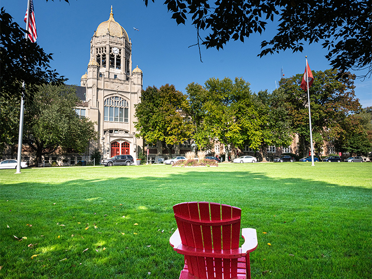 A red Adirondack chair sits beneath a tree with the clocktower of Haas College Center visible in the distance.