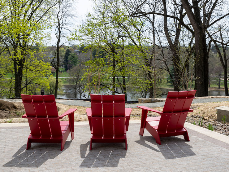 Three red Adirondack chairs sit on an outdoor stone plaza, facing a wooded space.