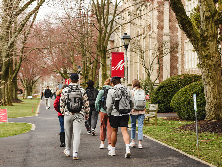 A group of students wearing backpacks walk away from the camera down Academic Row on the Muhlenberg College campus.