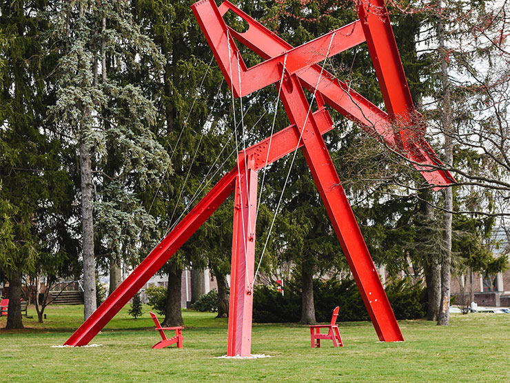 Two red Adirondak chairs sit beneath the red, modern sculpture, Victors Lament on the campus of Muhlenberg College.