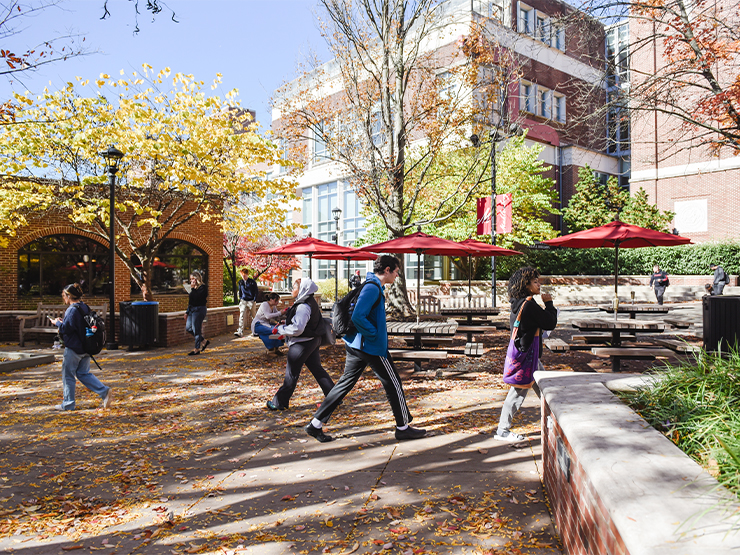 As autumn leaves fall from trees, two students walk to the right and two to the left in front of a plaza with tables topped with red umbrellas.