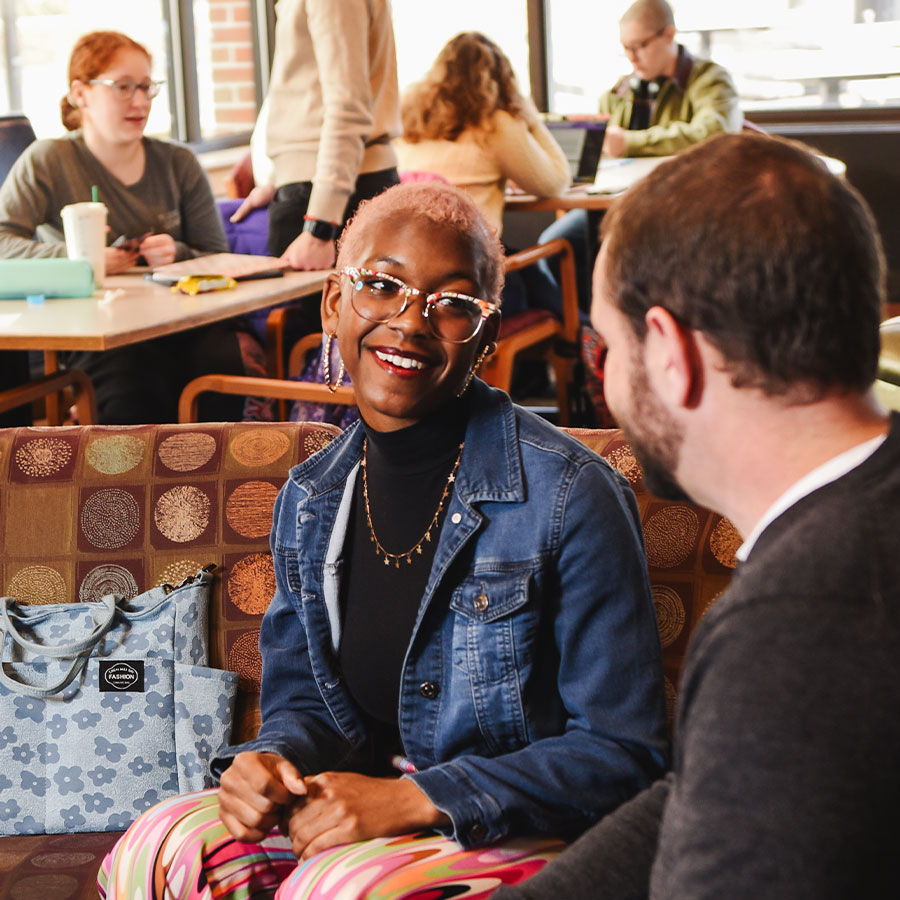 A student wearing a black turtleneck and denim jacket sits on a coach in a common area and smiles while speaking with an adult.