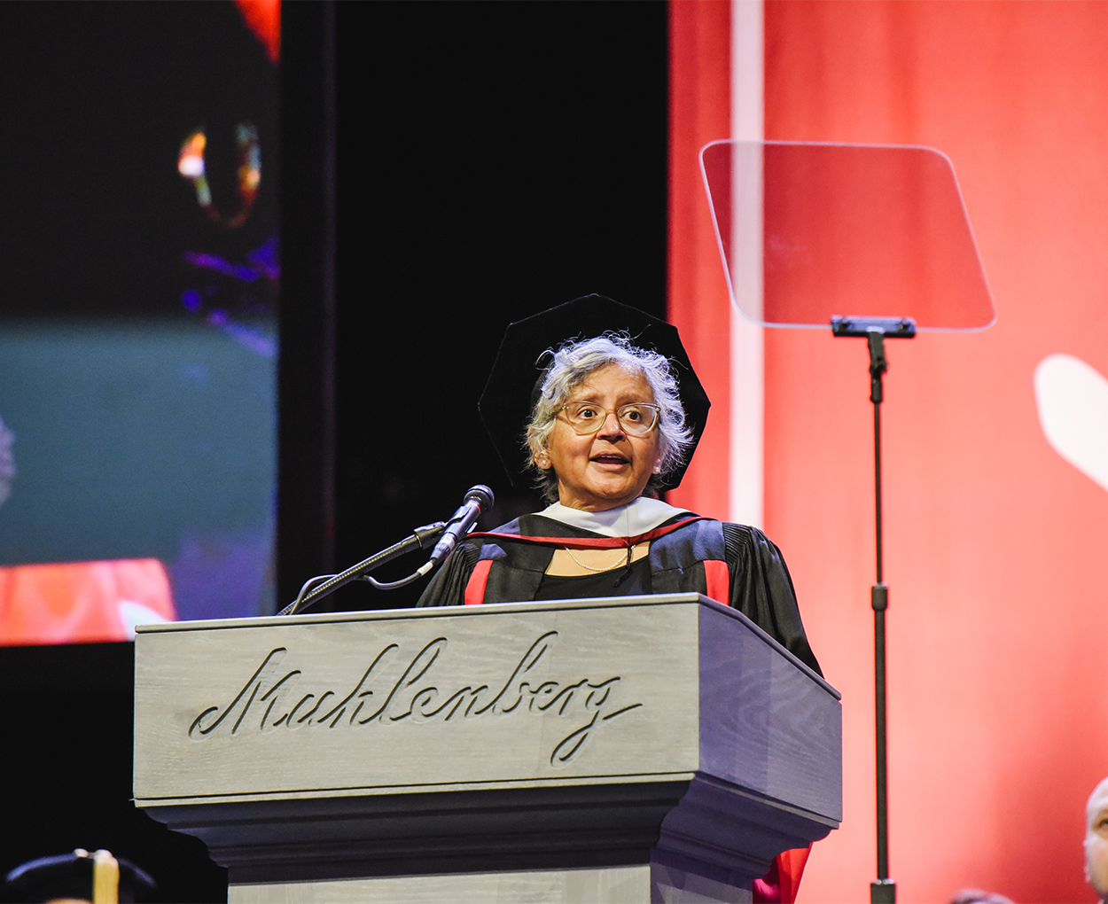 A woman in full commencement regalia speaks on a stage at a podium.