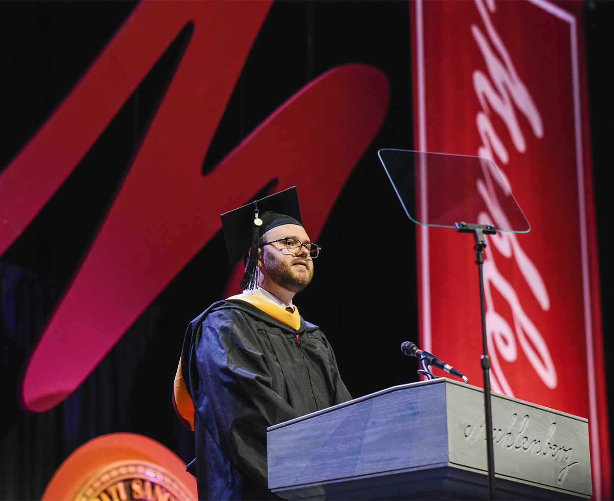 A male student in commencement regalia speaks at a podium.
