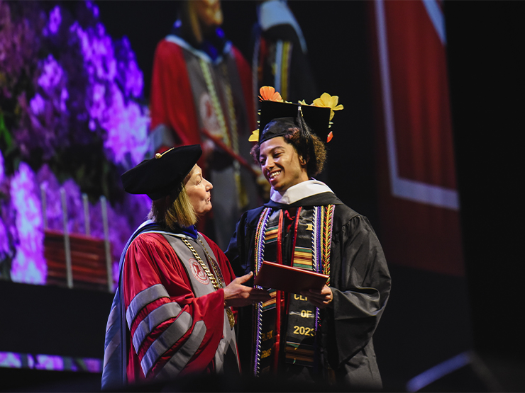 A woman in colligate president regalia congratulates a student in formal graduation regalia, with colorful flowers attached to the cap.