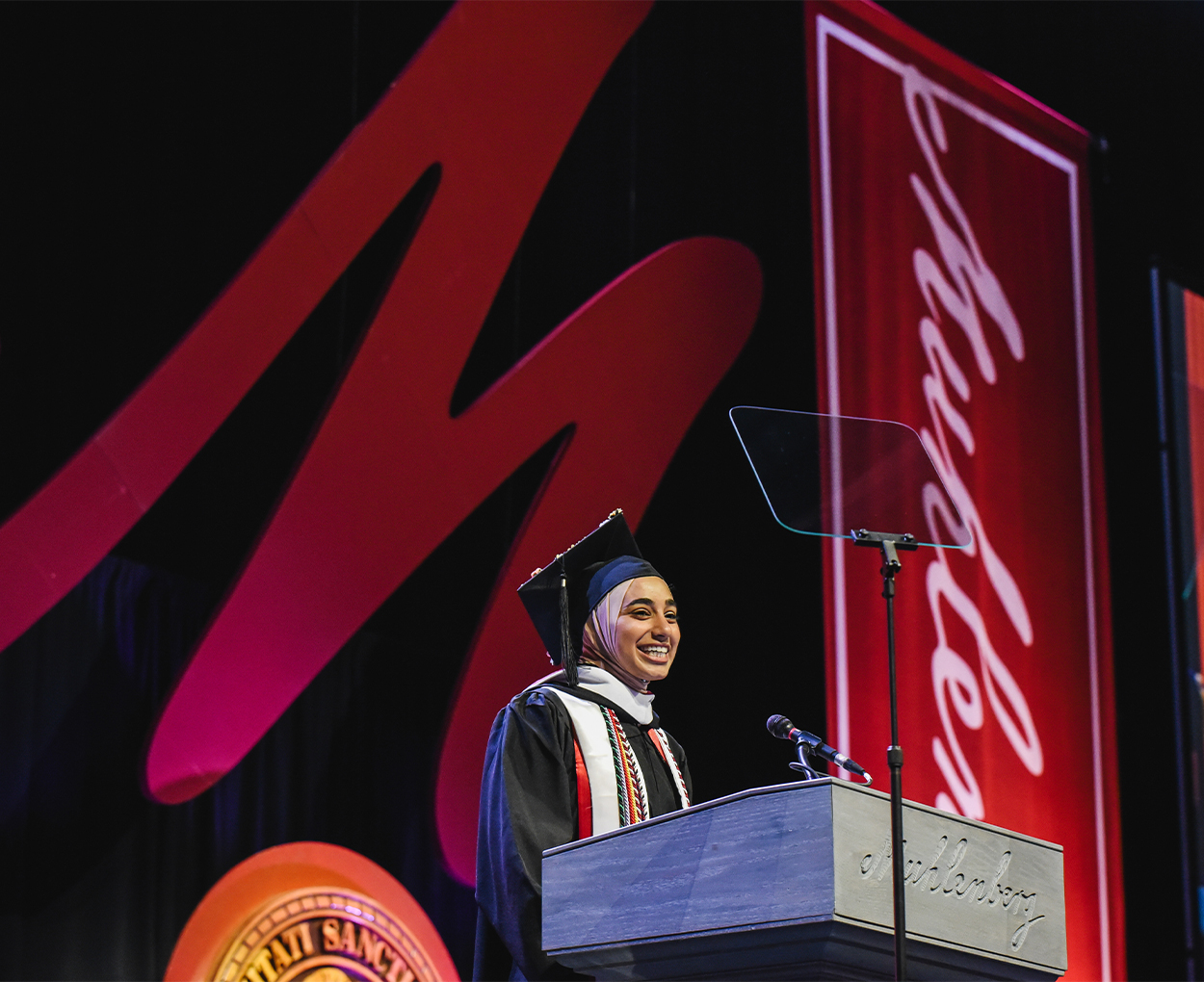 A female speaker with a headscarf is dressed in full commencement regalia is speaking at a podium on stage.