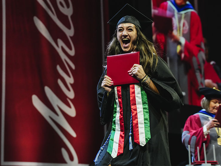 A student in graduation attire shouts in excitement while holding a diploma and making their way across a stage.