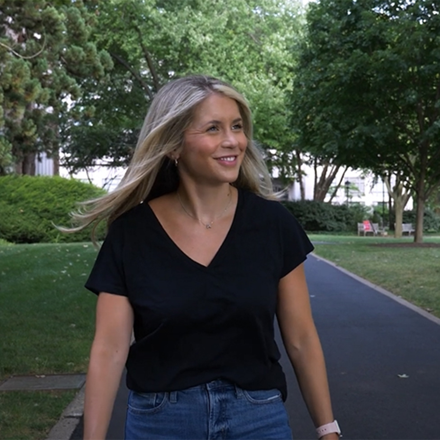 A woman with blonde hair and a black shirt walks along a sidewalk on a college campus.