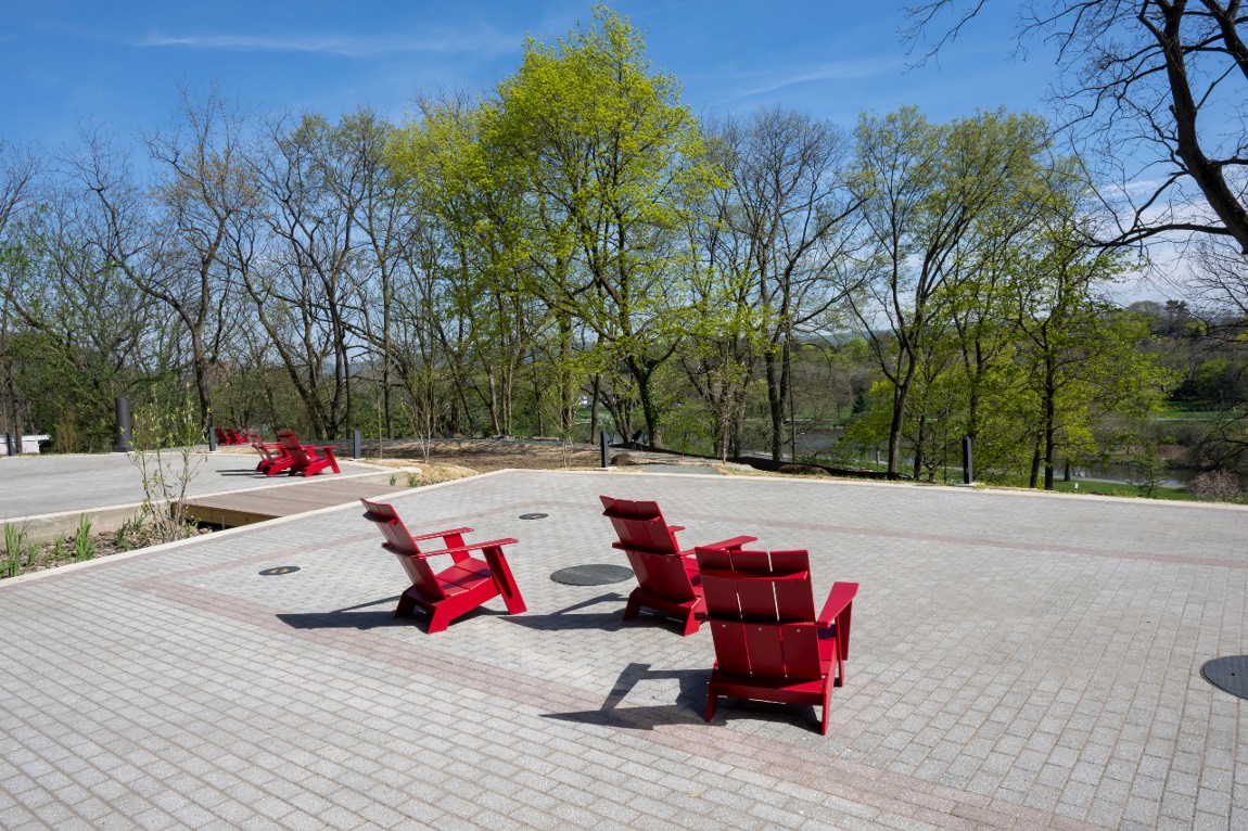 Large, sunny terrace with several red adirondeck chairs, on a hill overlooking trees and greenery.