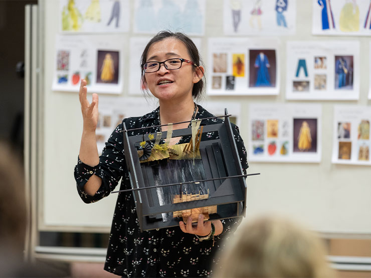 An adult instructor holds a model of a stage, behind them is a whiteboard covered in printed papers showing costume designs.