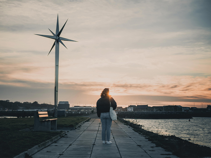 A woman stands on a sidewalk along a river, gazing at the setting sun. A tall sculpture towers in the distance.