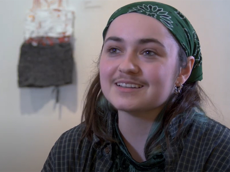 A young adult wearing a green bandana in their hair speaks to the camera in an art gallery.
