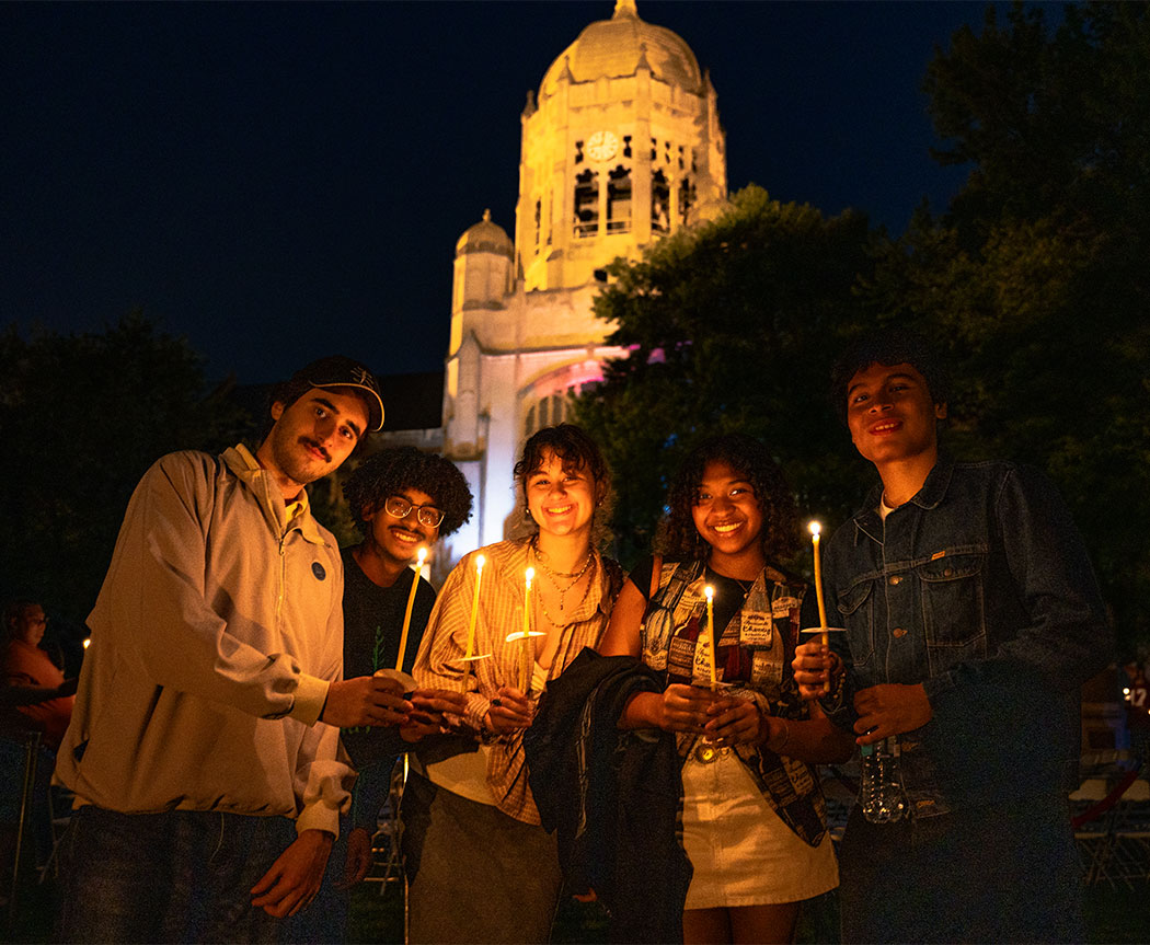 A group of four college students hold candles and smile with the Haas College Center lit against the dark sky behind them.