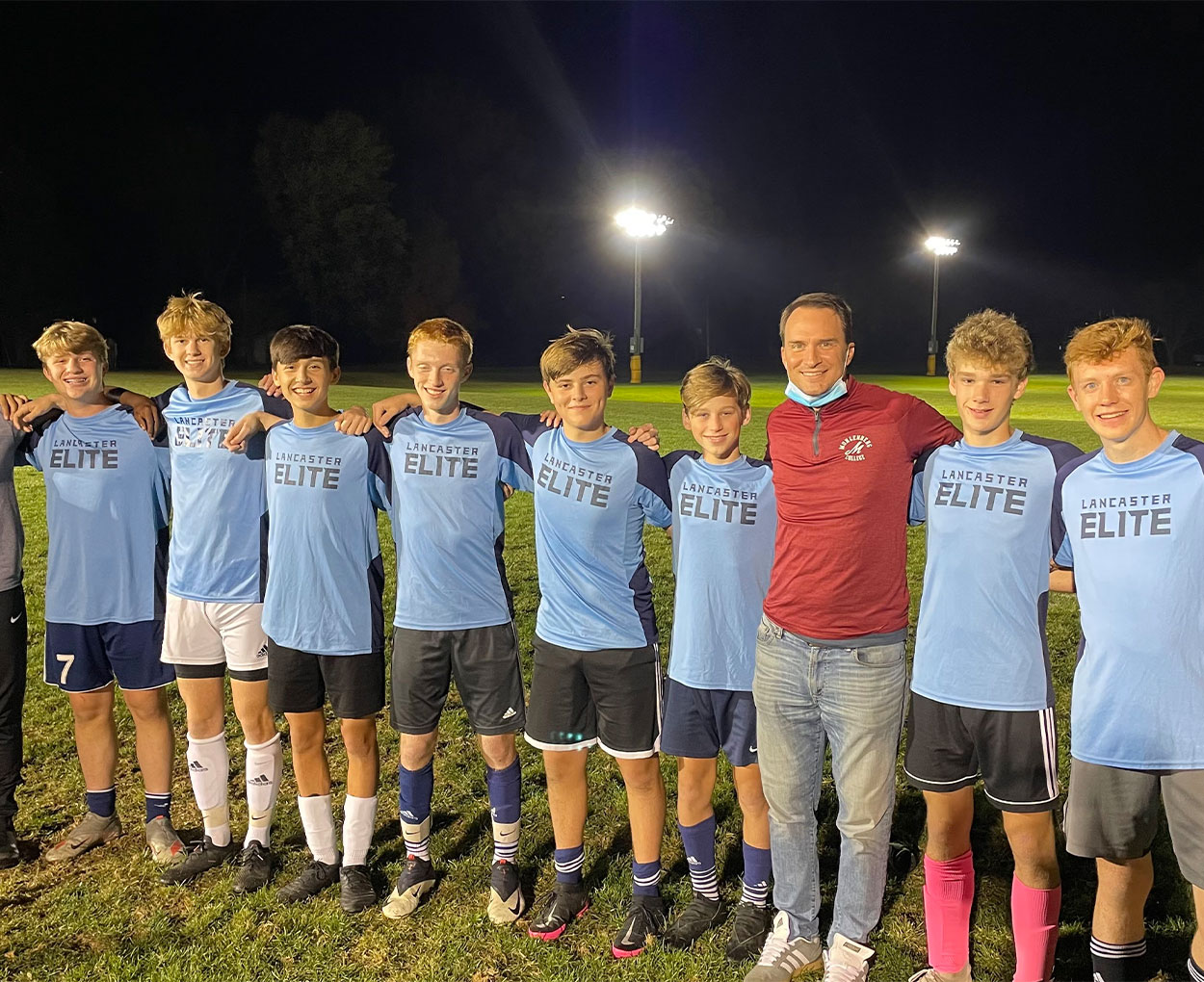 An adult in a red shirt poses with several teenage athletes in light blue jerseys.