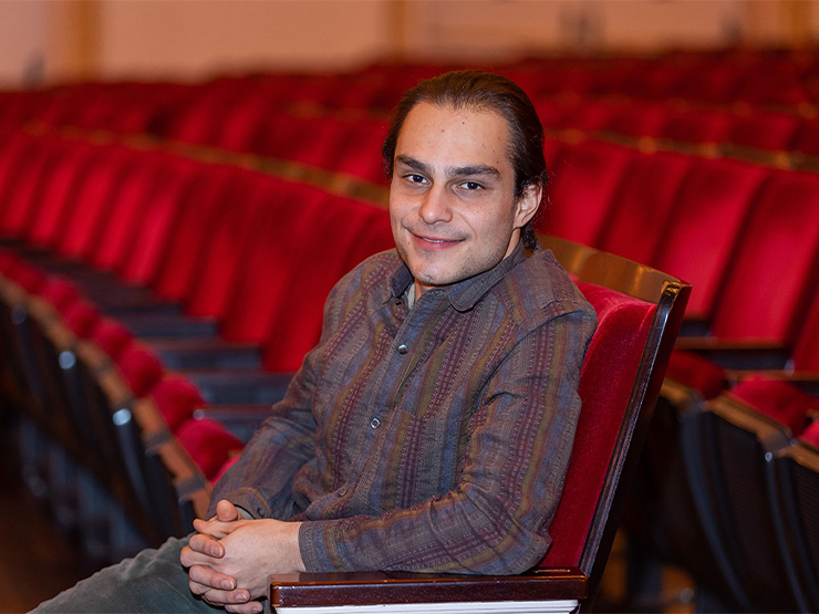 A man is seated, alone, in a concert hall filled with red velvet chairs.