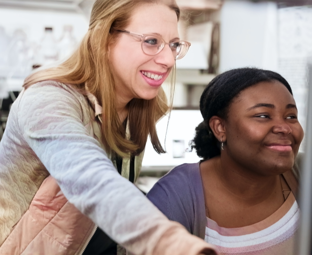 A chemistry instructor leans over the shoulder of a student as they work on a research project in a lab.