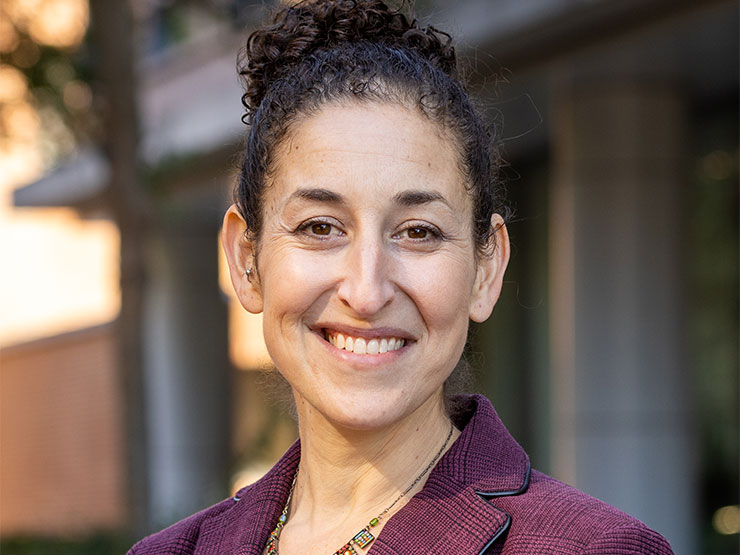 An adult, hair pulled back and wearing a purple blazer, smiles at the camera.