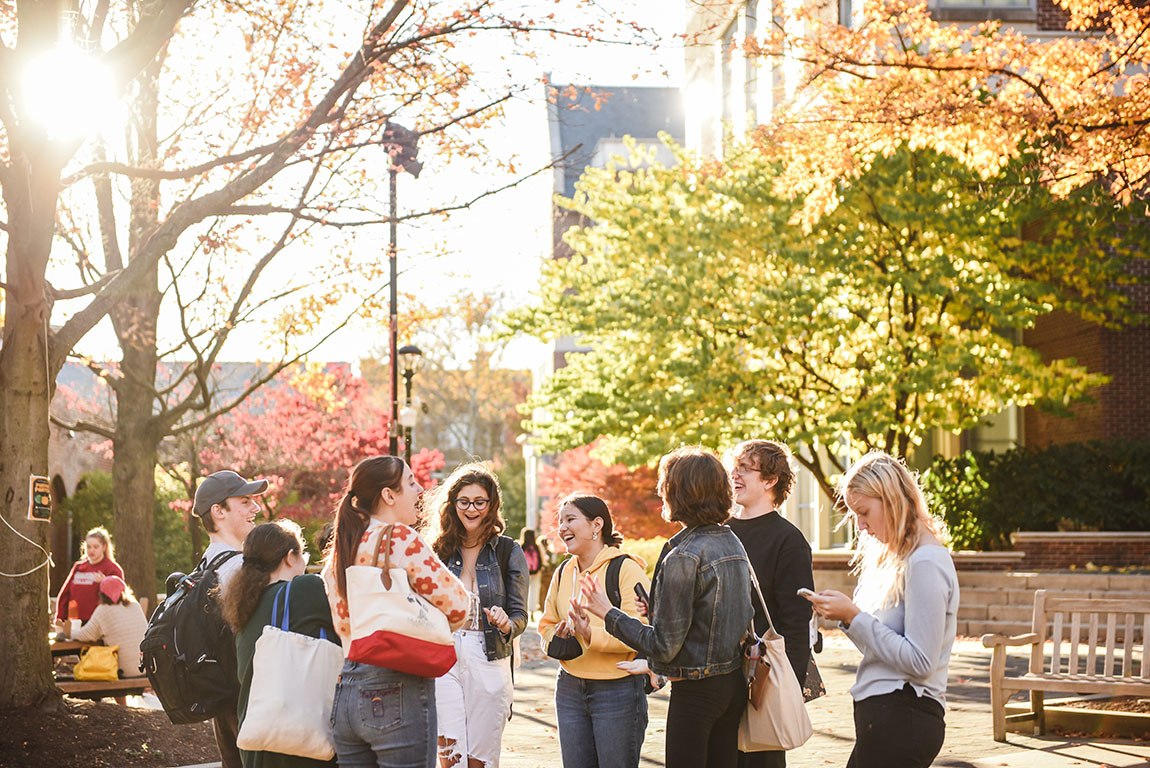 A group of college students stands in a circle, smiling and laughing, outside in the fall