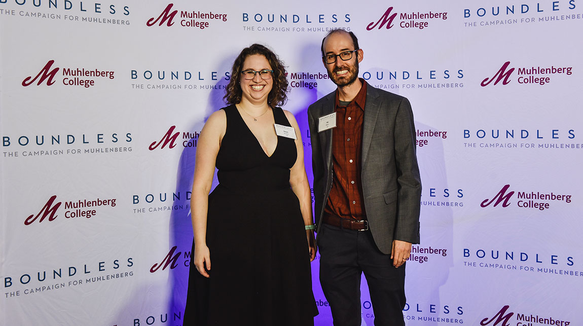 A woman in a black dress wearing glasses and a man in a suit with a beard wearing glasses smile in front of a backdrop that says Boundless: the Campaign for Muhlenberg