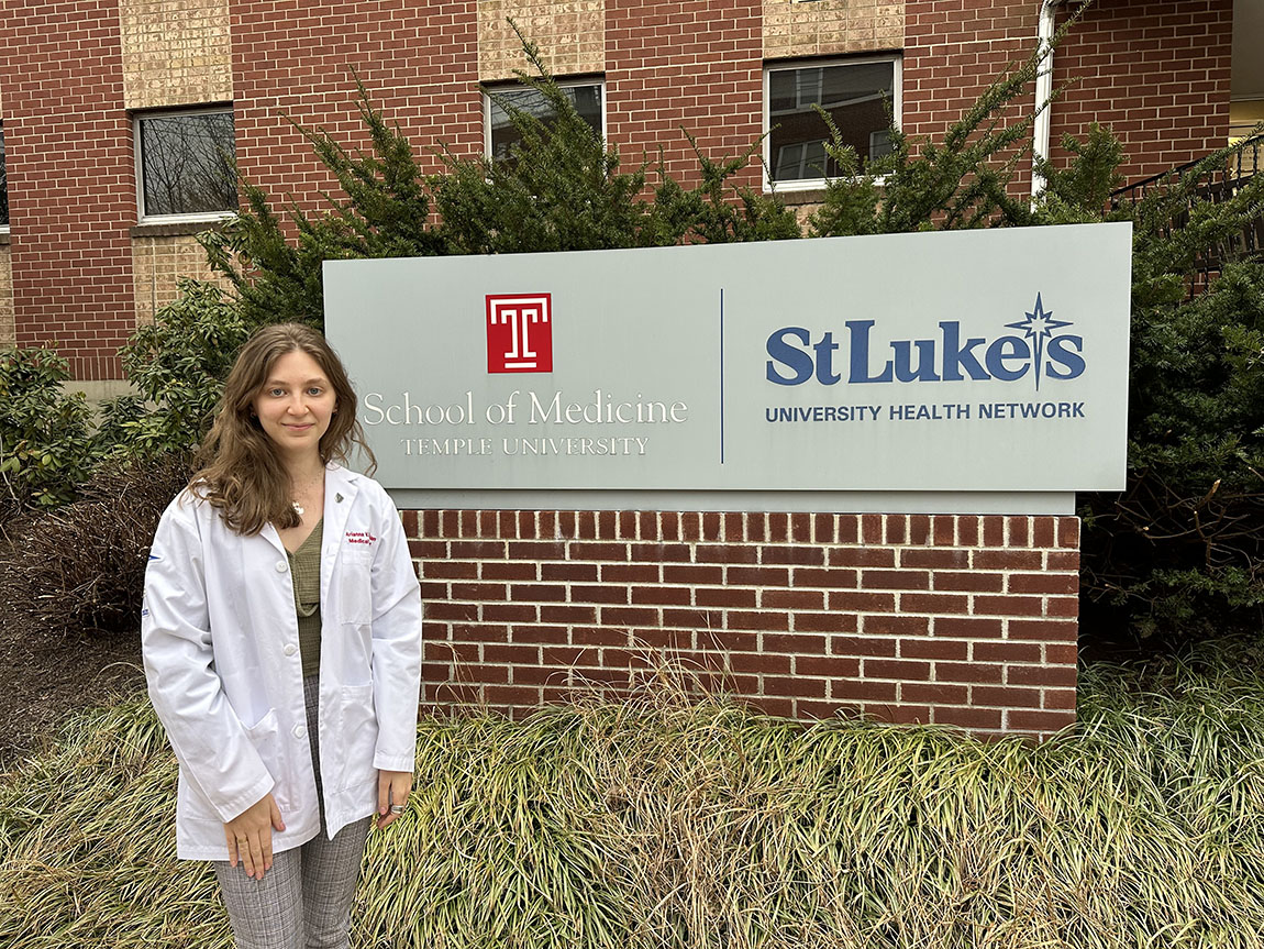 A young woman in a white doctor's coat stands in front of the sign for the Temple/St. Luke's medical school