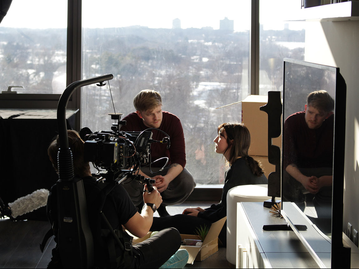 Two actors perform in front of a video camera with a big window overlooking a city behind them