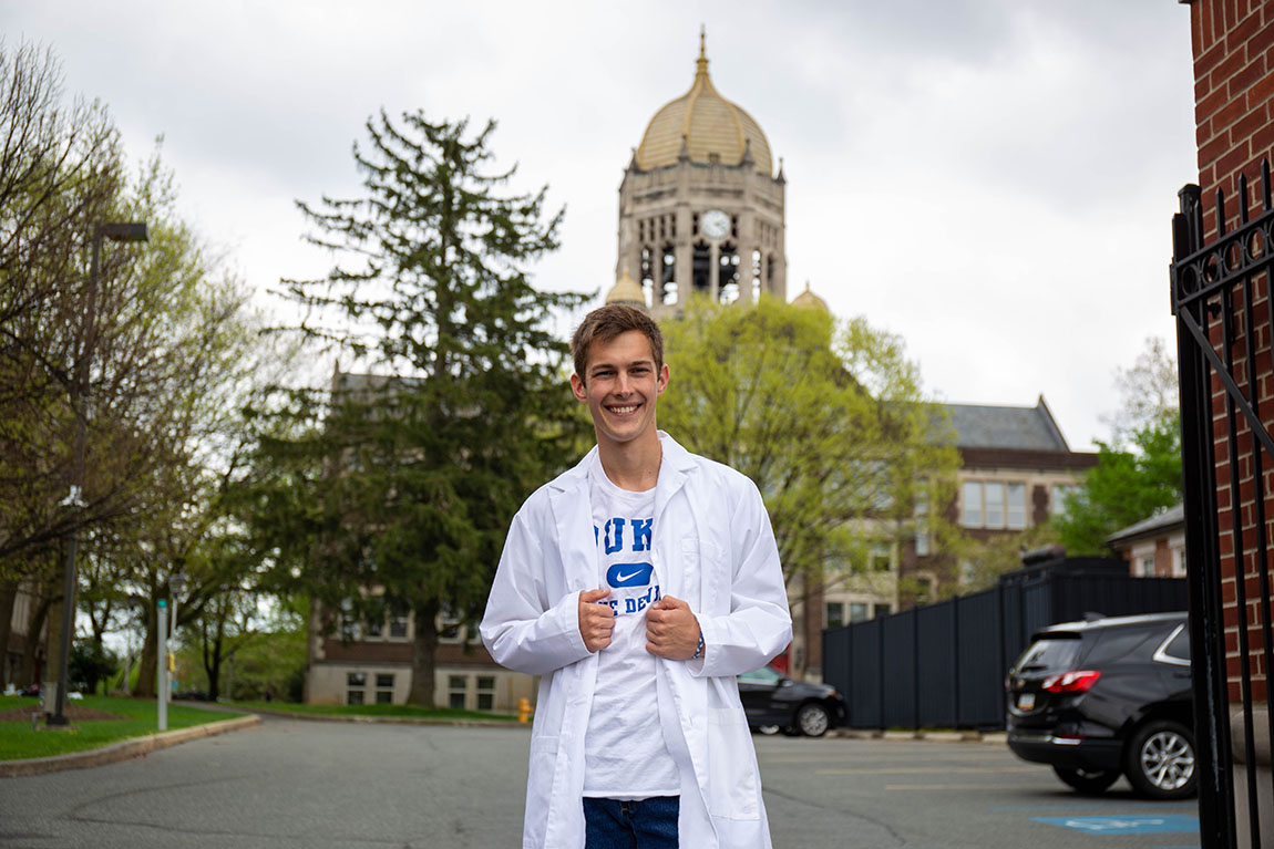 A white man with brown hair standing in front of the Haas Bell Tower in a white lab coat over a white Duke T-shirt