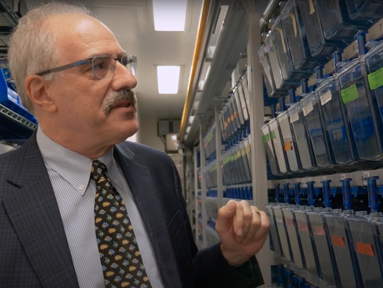 A scientist in a suit and tie stands in a room full of fish tanks