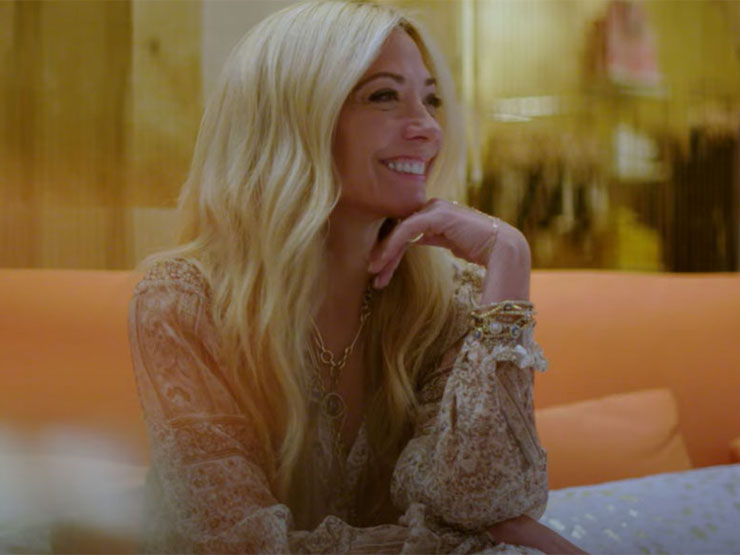 An adult with long blonde hair sits with a hand under the chin inside a clothing store, wearing a flowy tan dress with tiny details and jewelry.