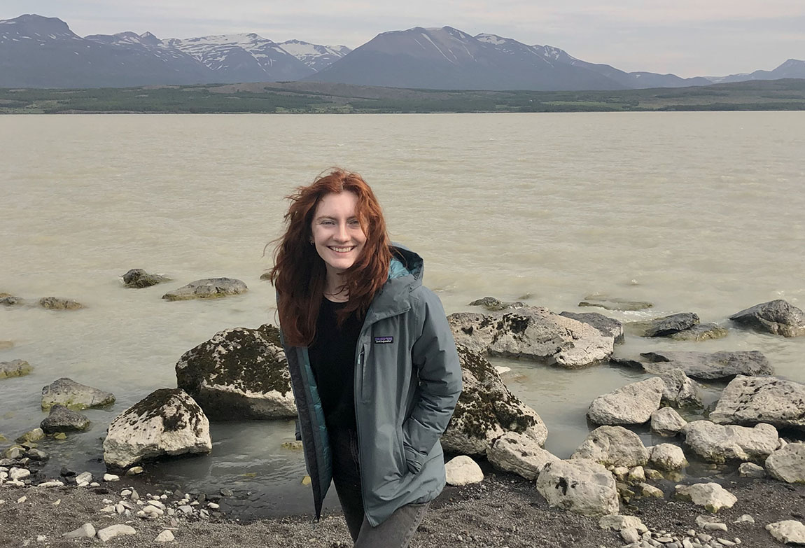 A smiling young adult with long red hair stands in front of rocks, water, and mountains