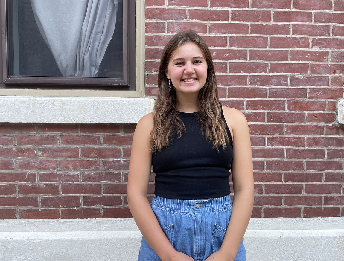 A white woman with brown hair stands, smiling, in front of a brick wall