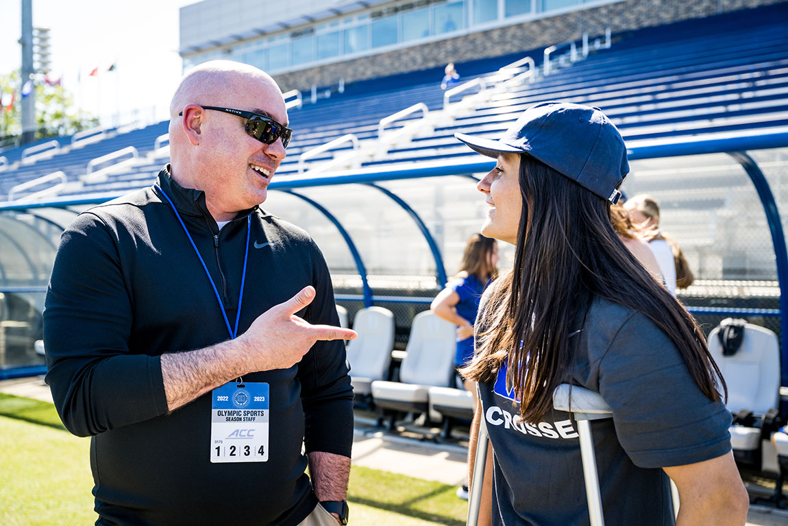 A bald man in sunglasses speaks with a college student with long hair wearing a baseball cap and using crutches with bleachers in the background