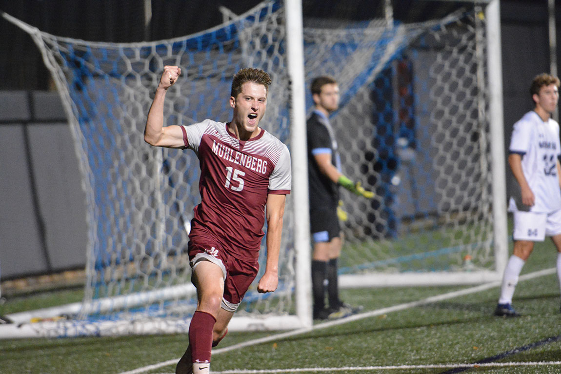 A college student in a soccer uniform on a field pumps his fist in celebration