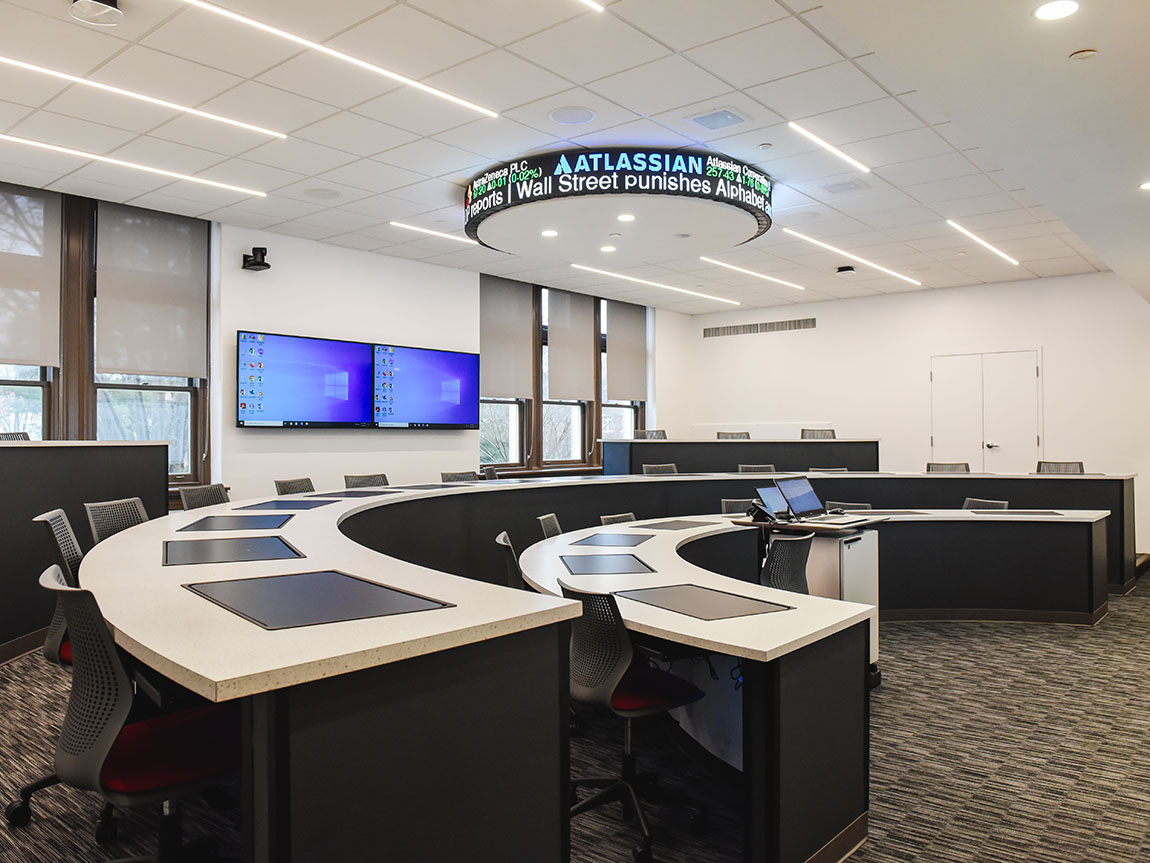 An empty classroom with stadium seating, big screens in the back and a stock ticker on the ceiling