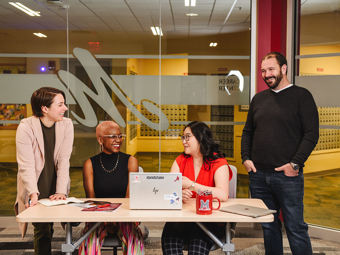 Three college faculty/staff and one student sit and stand around a table