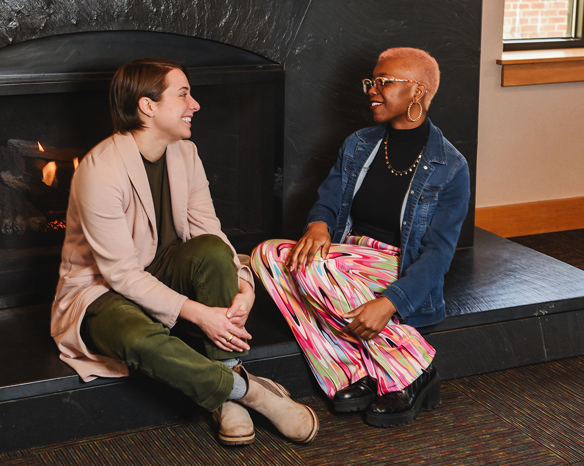 A college staff member sits with a college student on the hearth of a fireplace