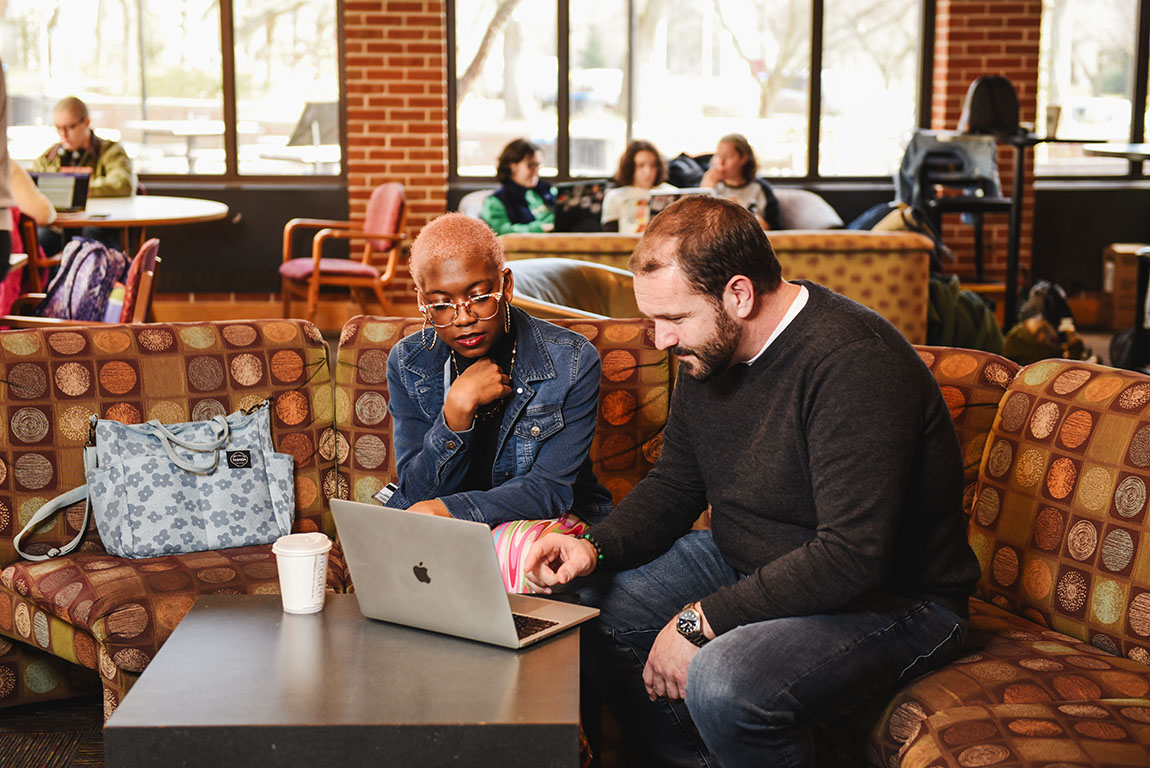 A college student sits with a faculty member and looks at a laptop