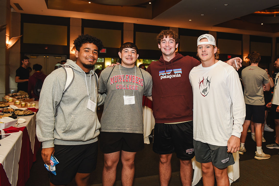 Four smiling college students pose for a photo inside a large, crowded room