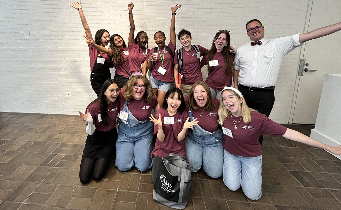 A group of 11 college students in matching maroon T-shirts and one staff member in a white button down and bow tie smile and reach their arms out