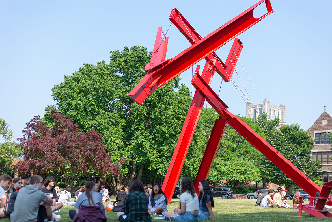 A large red sculpture made of steel beams in front of trees and a blue sky; groups of college students gather on the grass around the sculpture