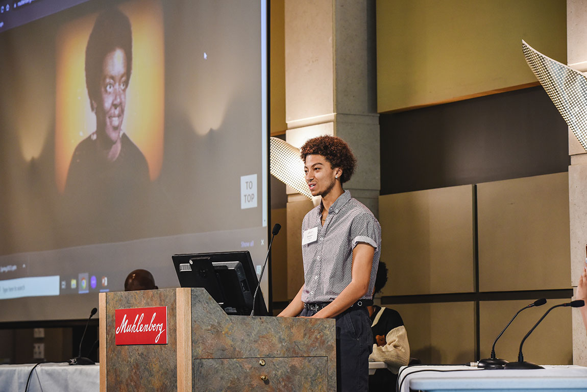 A college student in a short-sleeved button down shirt speaks at a podium with a picture of a woman projected on the screen behind him