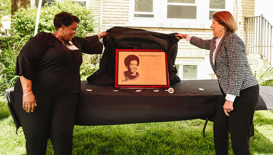 Two women unveil a bronze plaque with a photo of another woman on it and words next to her picture