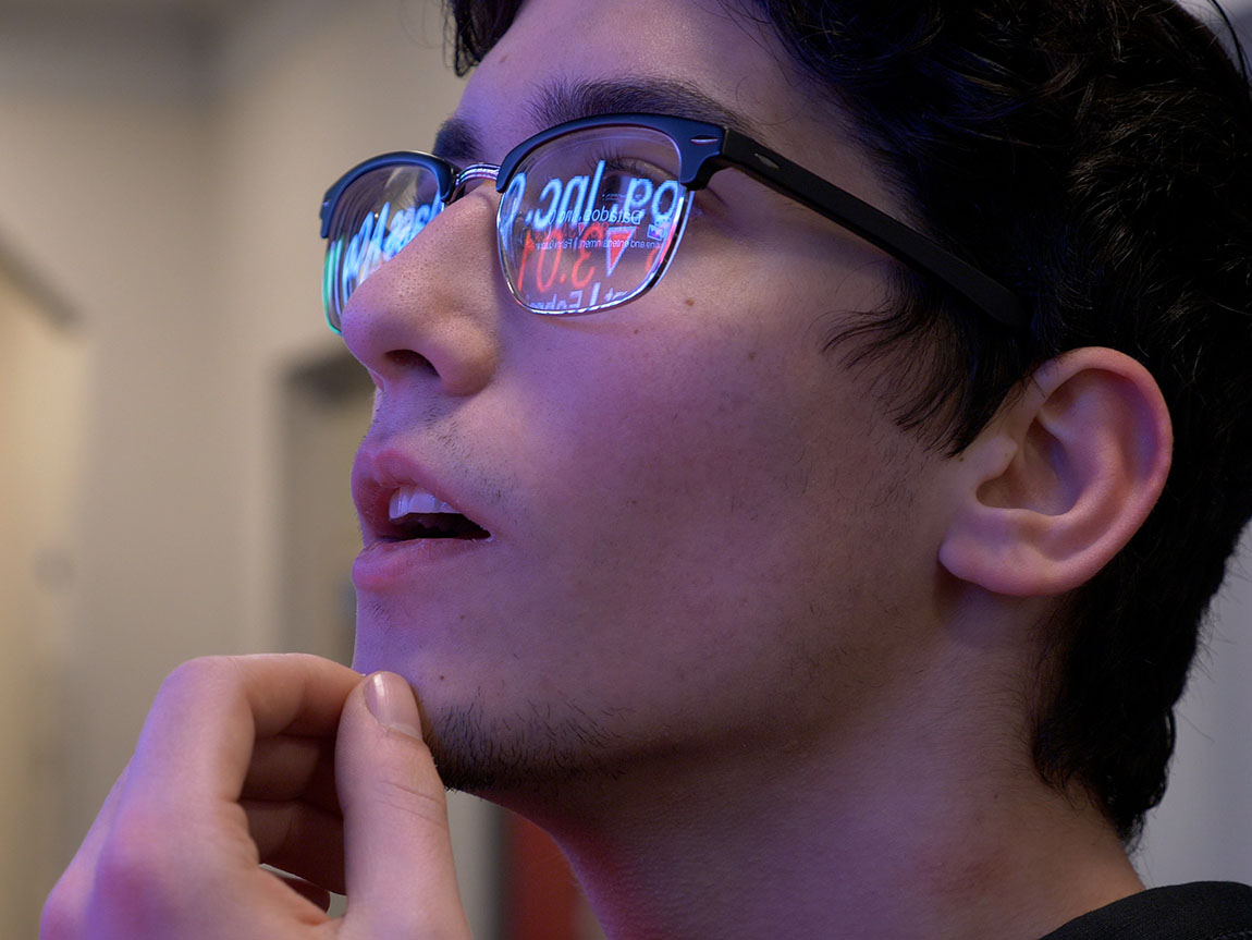 A college student wearing glasses looks at a screen displaying stock information that's reflected in his glasses