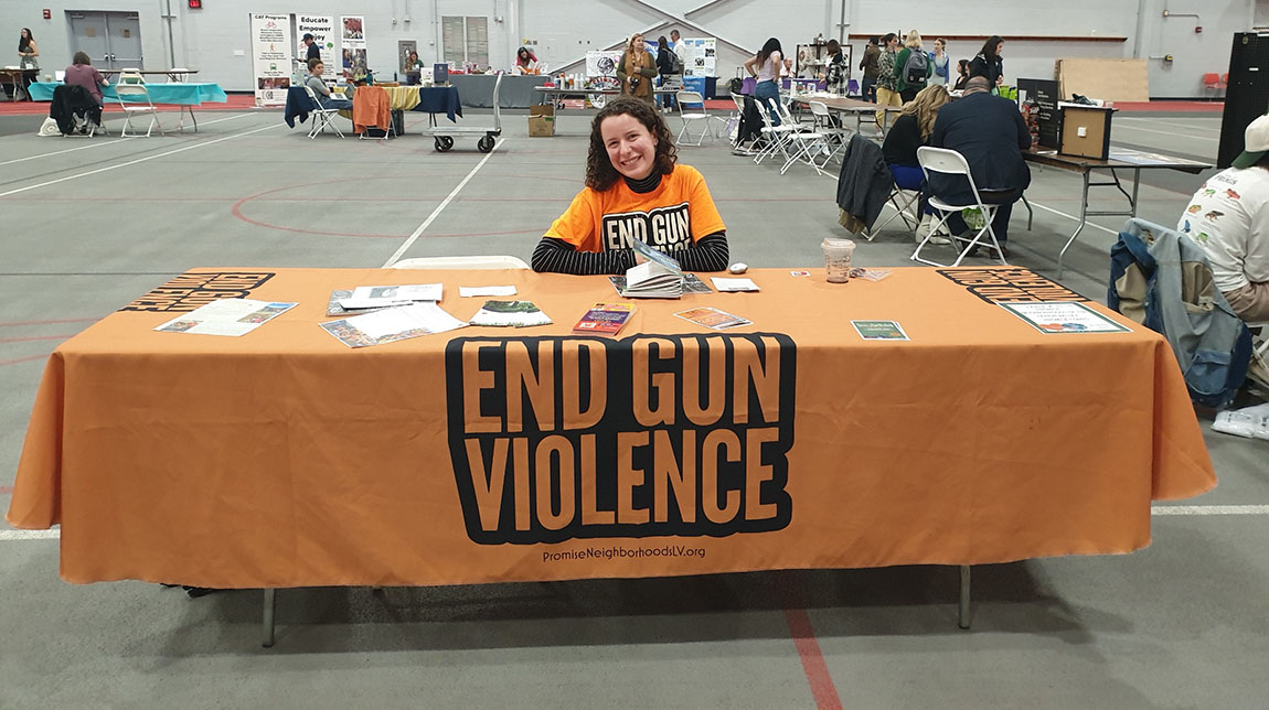 A smiling college student sits at a table in a large indoor space that's covered by an orange tablecloth that says End Gun Violence