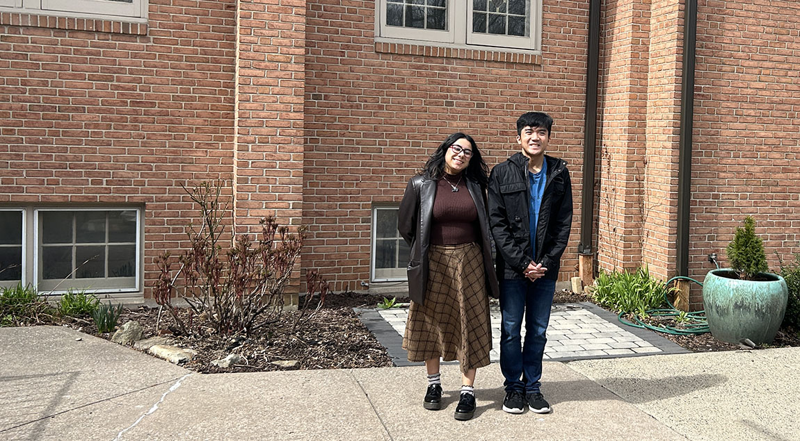 Two college students stand smiling on the sidewalk outside a brick building