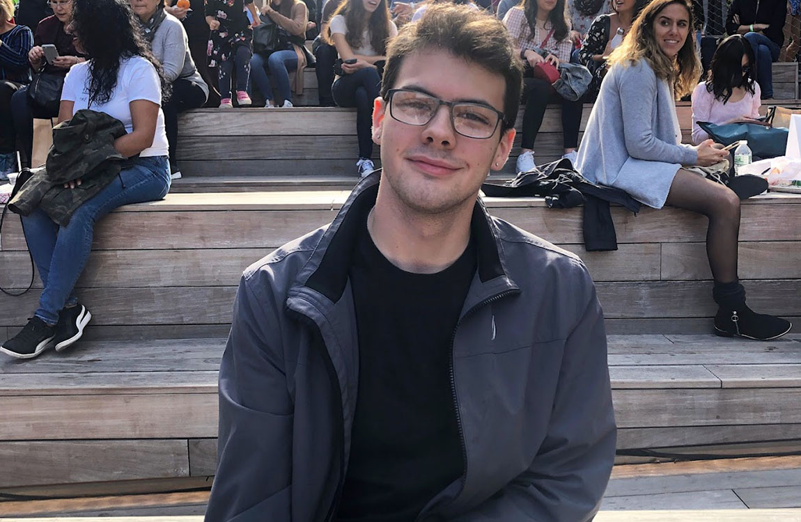A young man with dark hair and glasses in a black shirt and jacket smiles at the camera while sitting on some bleachers