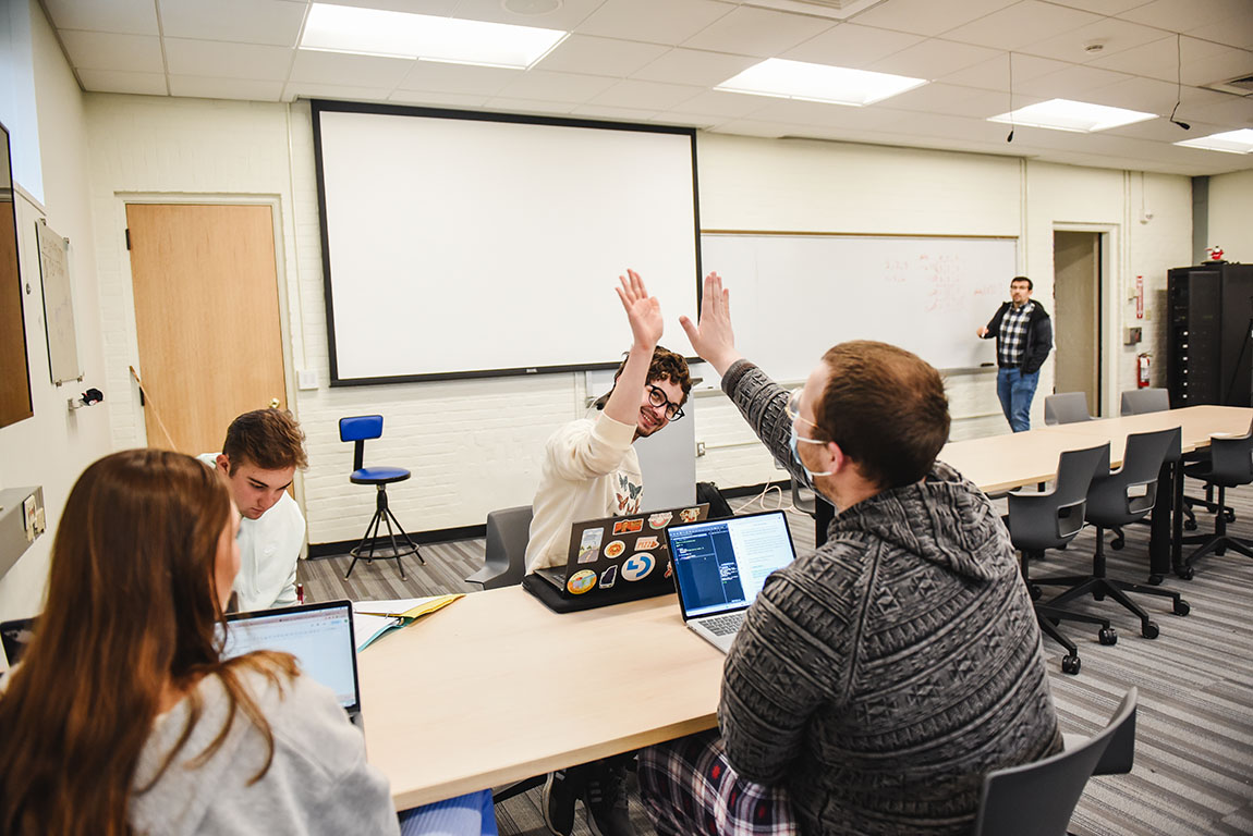 Two college students high-five each other in class while their professor looks on in the background